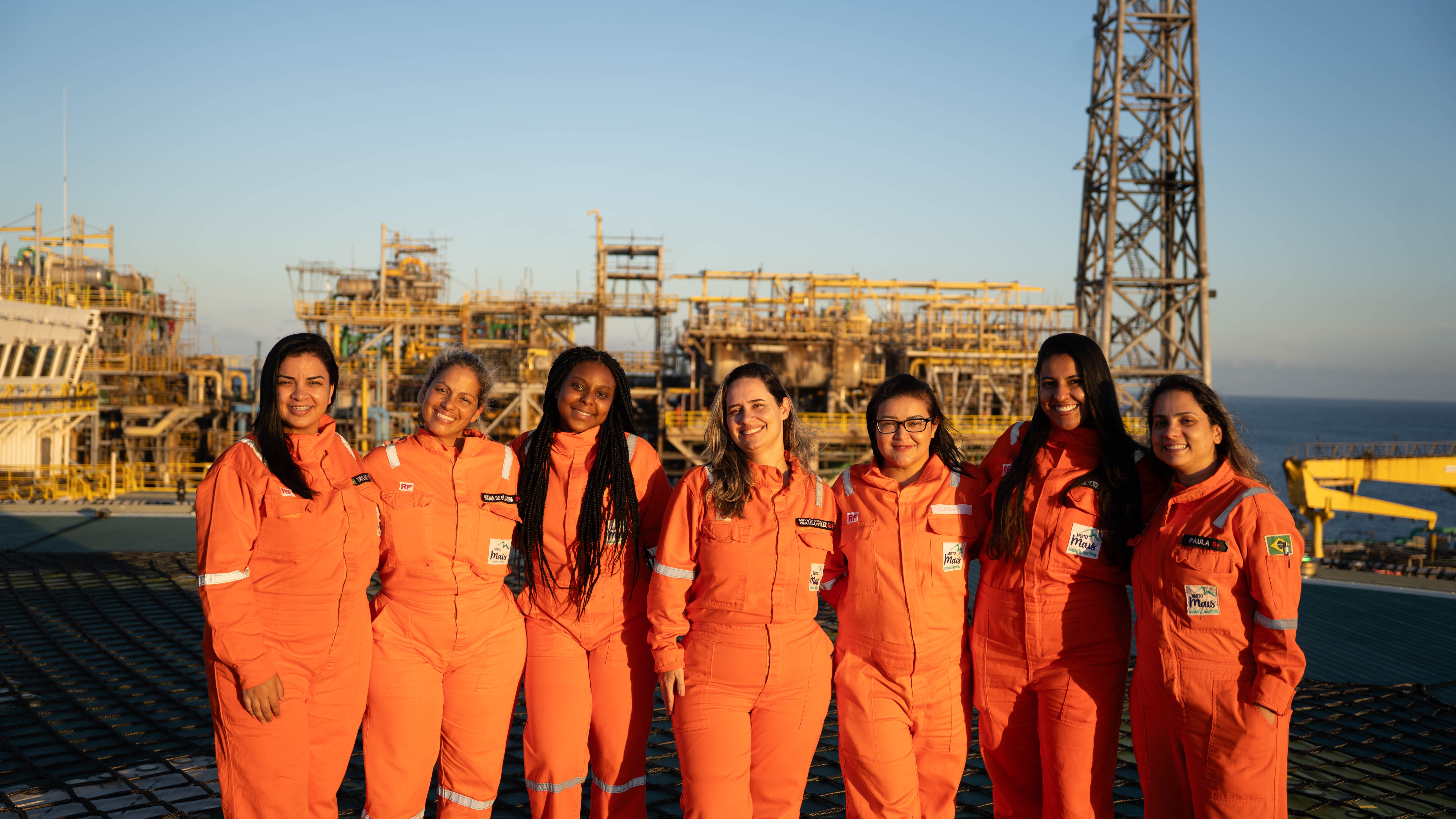 Group of women in orange work uniform standing at an industrial site with machinery in background